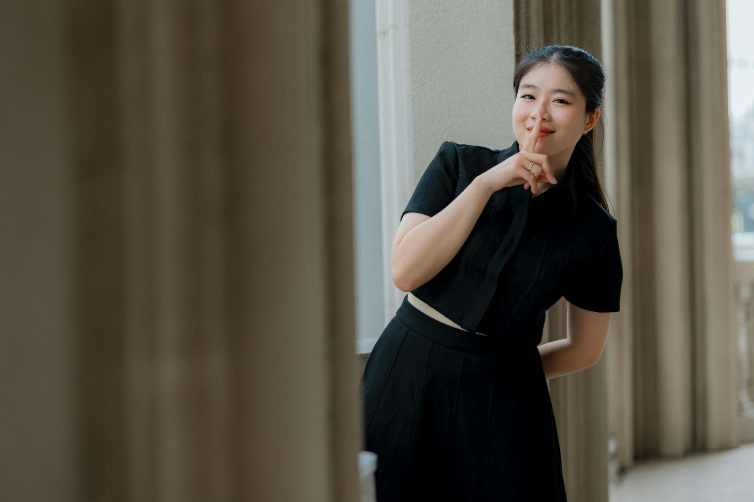 Young woman in black dress indoors making a silence gesture by her lips.