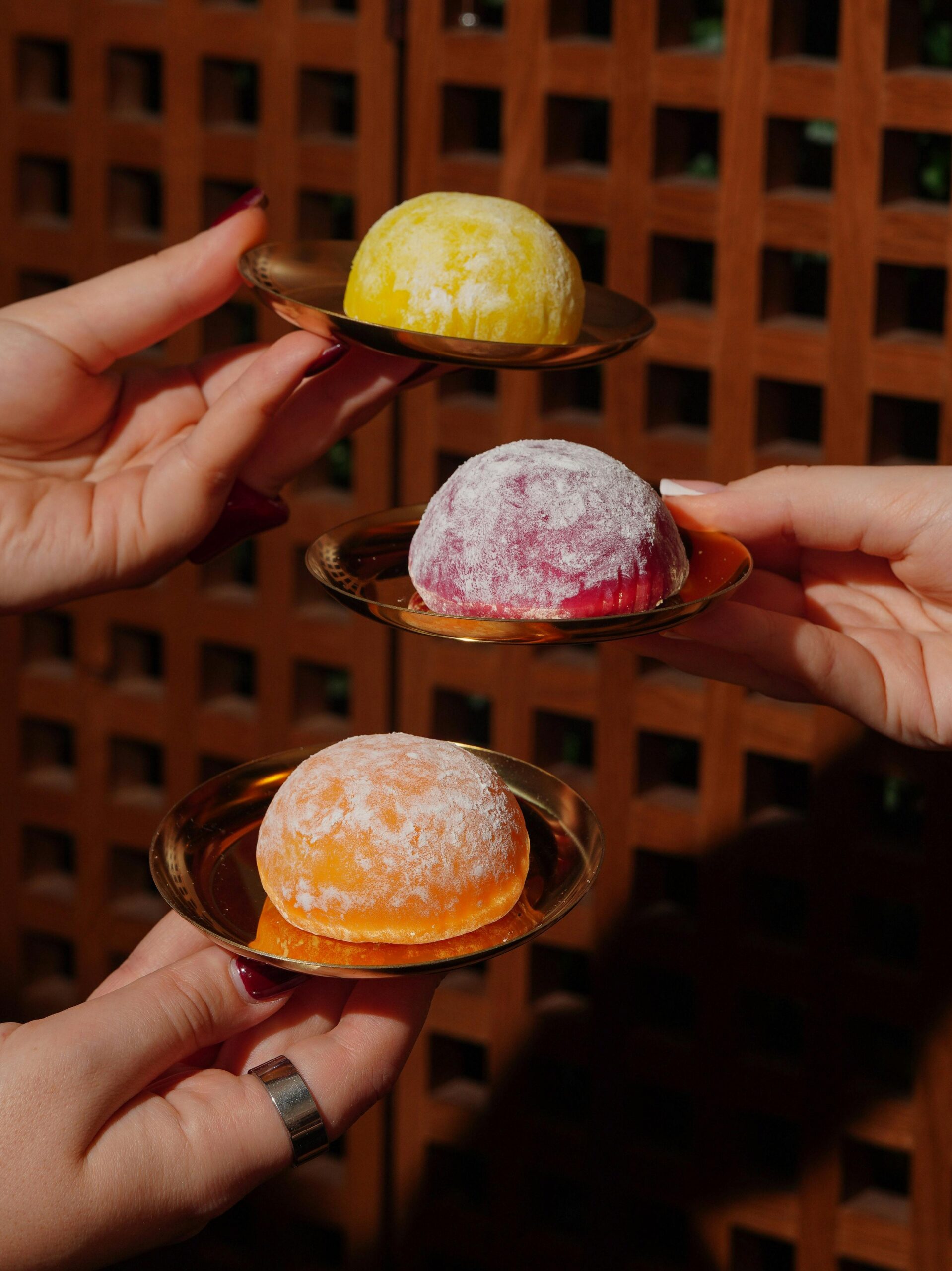 Three colorful mochi desserts presented on plates, held by hands outdoors.